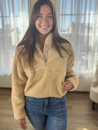 Woman wearing a beige fleece pullover and blue jeans indoors with large windows in the background