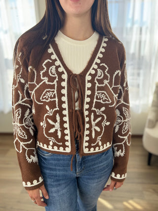 Person wearing a brown cardigan with white patterns, white shirt, and blue jeans indoors.
