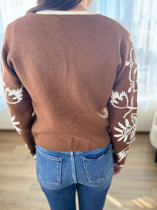 Brown sweater with white floral patterns worn by a person, standing indoors.