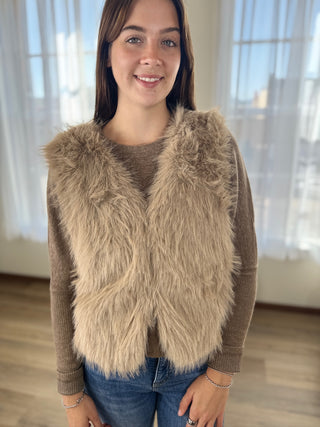 Woman wearing a brown sweater with a beige fur vest indoors.