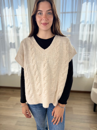 Woman wearing a beige cable knit sweater over a black shirt with a blurred indoor background