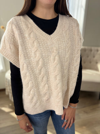 Person wearing a beige cable knit sweater vest over a black shirt with a blurred indoor background