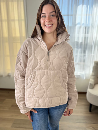 Woman wearing a beige quilted pullover in a room with large windows.