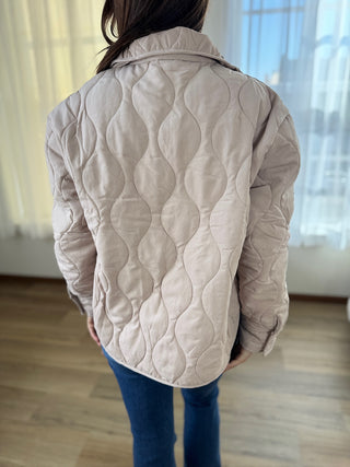 Person wearing a beige quilted jacket in a room with light-colored walls and wooden floor.
