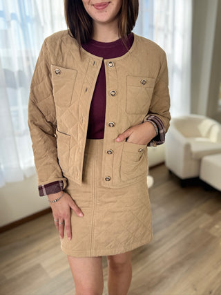 Woman wearing a beige jacket and skirt in a room with white curtains and furniture.