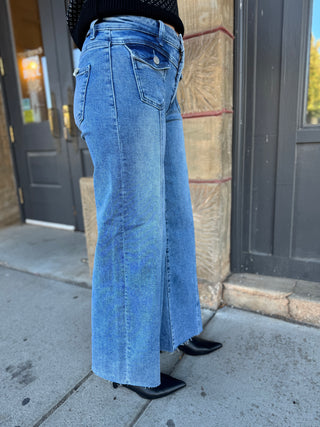 A person wearing light blue wide-leg denim jeans with a raw hem, paired with black pointed-toe heels, standing on a sidewalk in front of a building entrance.