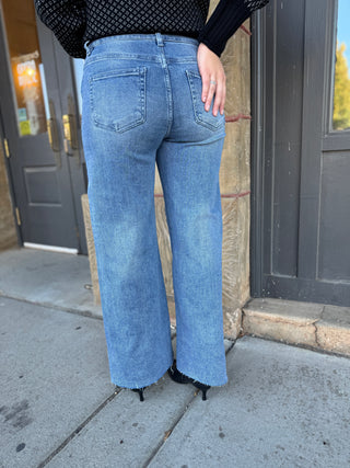 A person shown from the back wearing light-wash blue wide-leg jeans with a frayed hem, paired with a black patterned top and black heels, standing outdoors on a sidewalk in front of dark storefront doors.
