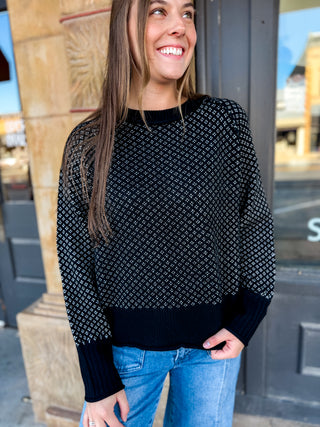 A woman wearing a black knit sweater with a white geometric pattern and ribbed cuffs and hem, paired with blue jeans, standing outdoors in front of a storefront.
