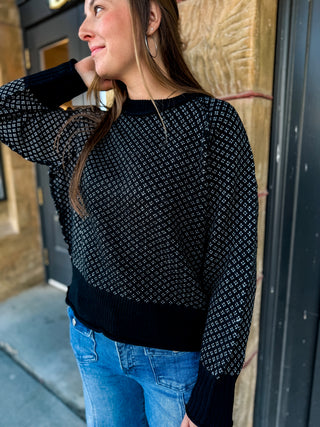 A woman wearing a black knit sweater with a small white geometric pattern, paired with blue jeans, standing outdoors near a building entrance.