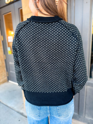 A person viewed from the back wearing a black knit sweater with a white geometric lattice pattern, paired with light blue jeans, standing outdoors in front of a gray door.