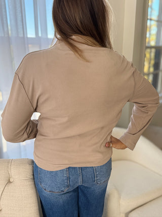 Person wearing a beige long-sleeve top and blue jeans in a room with a couch and window.