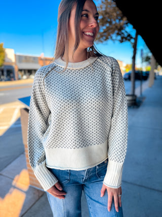 A woman wearing a cream and gray geometric patterned knit sweater with blue jeans, standing outdoors on a sunny sidewalk.