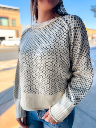 A woman wearing a cream and black patterned knit sweater with blue jeans, standing outdoors on a sunny day.