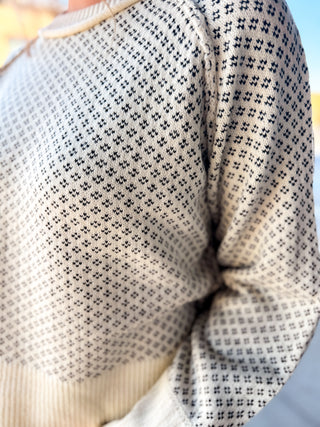 Close-up of a cream knit sweater with a repeating black geometric cross-like pattern, worn by a person.