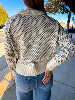 A person viewed from behind wearing a cream and black geometric patterned knit sweater with ribbed collar, cuffs, and hem, paired with blue jeans, outdoors on a sunny day.