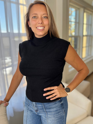 Woman wearing a black sleeveless turtleneck and blue jeans indoors.