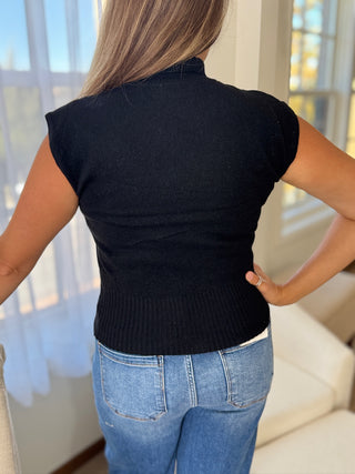 Person wearing a black sleeveless sweater and blue jeans indoors.