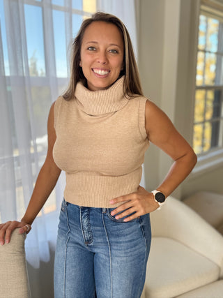 Woman wearing a beige sleeveless sweater and blue jeans indoors.