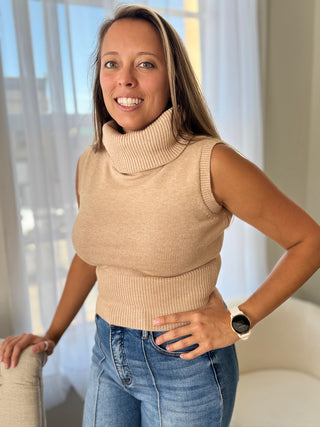 Woman wearing a beige sleeveless sweater and blue jeans indoors.