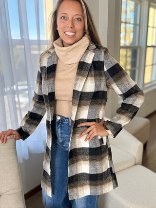 Woman wearing a plaid coat, beige sweater, and blue jeans indoors.