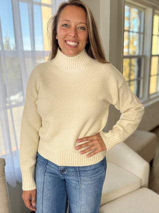 Woman wearing a cream sweater and blue jeans indoors with large windows in the background
