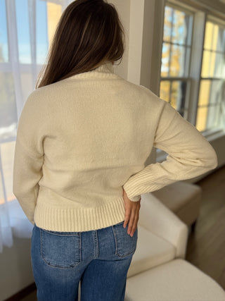 Person wearing a beige sweater and blue jeans standing in a room with large windows.