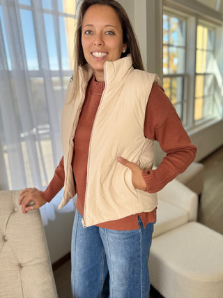 Woman wearing a beige puffer vest over a brown shirt and blue jeans in a living room.