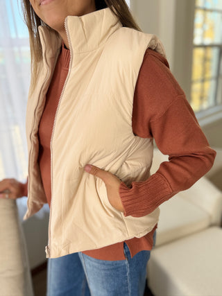 Person wearing a beige puffer vest over a brown long-sleeve shirt with blue jeans indoors.