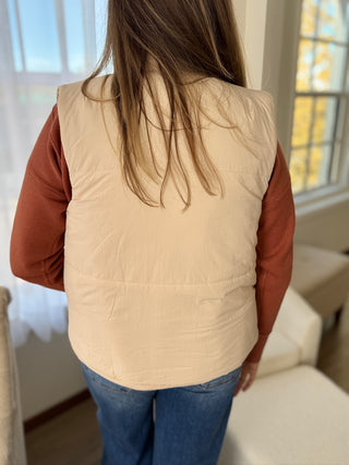 Person wearing a beige puffer vest over a brown shirt in a room with a window.