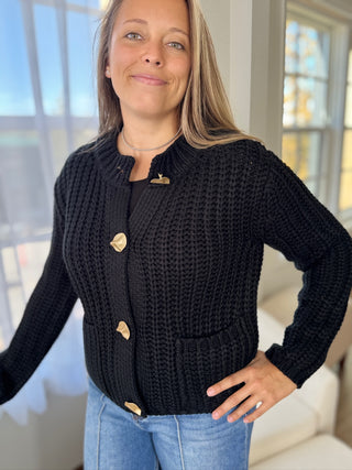 Woman wearing a black knitted cardigan with gold buttons indoors.