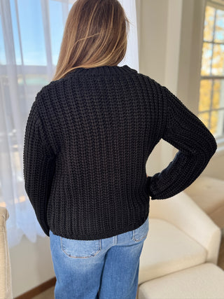 Person wearing a black sweater and blue jeans standing in a room with light-colored walls and furniture.
