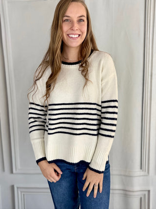 Woman wearing a cream sweater with black stripes and blue jeans against a white wall.