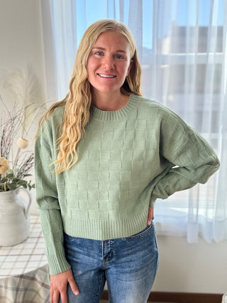 Woman wearing a green sweater and blue jeans indoors with a window in the background