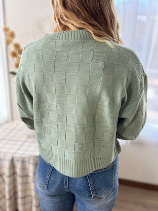 Person wearing a green textured sweater and blue jeans indoors.
