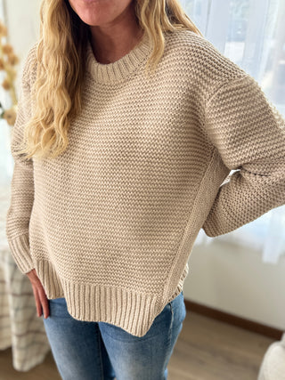 Person wearing a beige knitted sweater and blue jeans indoors.