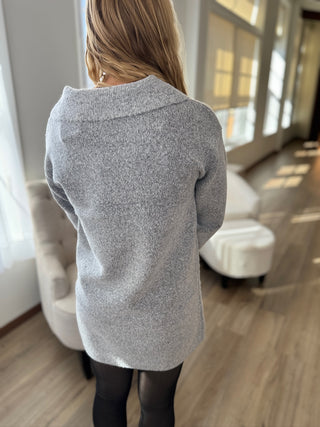 Person wearing a gray sweater in an indoor setting with chairs and a window.