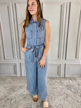 Woman wearing a denim jumpsuit standing against a gray paneled wall.