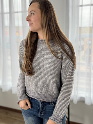 Woman wearing a gray sweater and jeans indoors with white curtains in the background