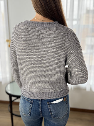 Person wearing a gray knitted sweater and blue jeans in a room with white curtains.