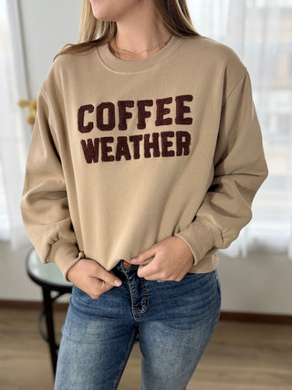 Person wearing a beige sweatshirt with 'COFFEE WEATHER' text, standing indoors.