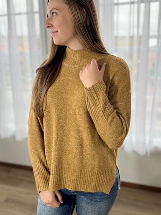 Woman wearing a mustard yellow sweater in a bright room with sheer curtains.