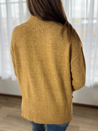 Person wearing a mustard yellow sweater in a room with light-colored curtains.