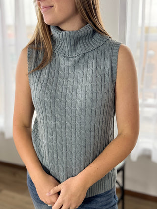 Person wearing a gray sleeveless knitted sweater in a bright room.
