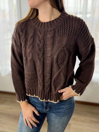 Person wearing a brown cable knit sweater and blue jeans indoors.