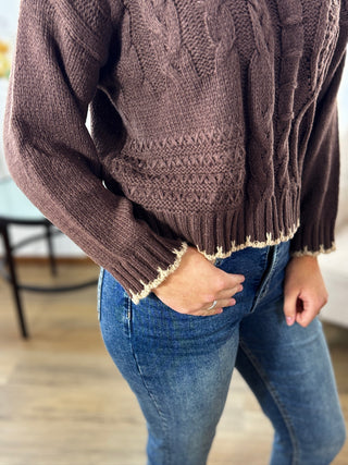 Person wearing a brown cable knit sweater and blue jeans indoors.