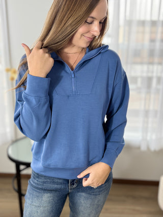 Woman wearing a blue pullover with a quarter zip in a room with a table and window blinds.