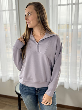 Woman wearing a light purple pullover with a quarter zip in a room with sheer curtains.