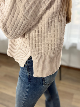 Person wearing a beige knitted sweater and blue jeans indoors.