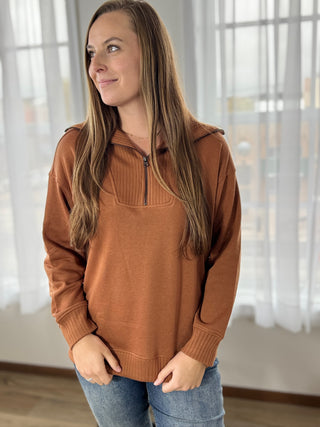 Woman wearing a brown sweater with a zipper in a room with sheer curtains.