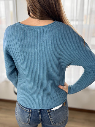 Person wearing a blue sweater and jeans indoors with a neutral background